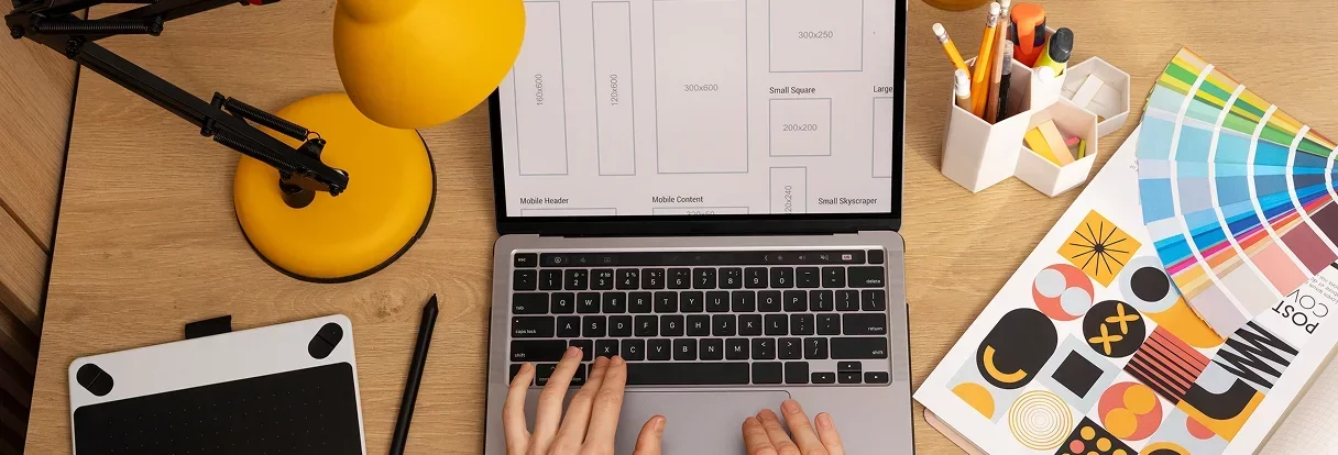 Person typing on a laptop keyboard on a wooden desk with a yellow desk lamp, graphic tablet and stylus, container with pencils and erasers, and color swatches with designs.
